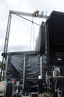 Der Seitenarm einer Festivalbühne (Eingang/Vorzelt) mit schwarzer Plane verkleidet. Darum ein ca. 7 bis 8 Meter hohes Gerüst, auf dem eine Person sitzt und einen Scheinwerfer montiert. Neben ihm hängt ein Sicherungsseil herunter, das von einer auf dem Boden stehenden Person mit Bauheln gehalten wird.