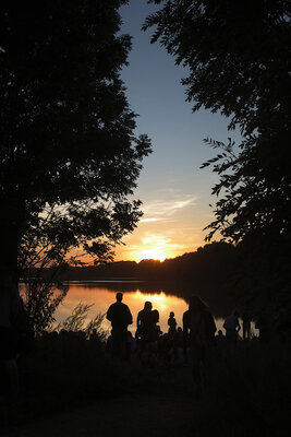 Ein Sonnenuntergang über einem großen, stillen See. Die Sonne und der große orangefarbene Fleck, den sie in den gerade noch blauen Himmel malt sind von Büumen umringt. Vor dem See stehen und sitzen ungefähr 25 Menschen und schauen sich die Szene an.