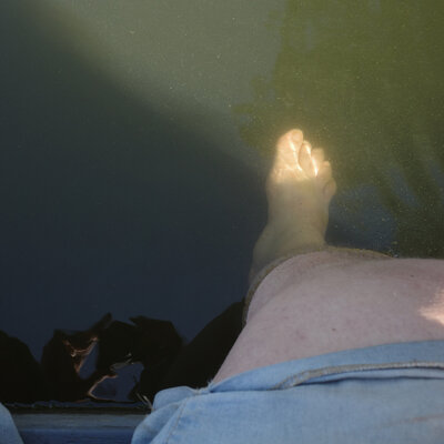 Ein nacktes Beim baumelt in grünlichem Wasser. Der Fuß wird von einem Sonnenstrahl zum gelb-grün-leuchten gebracht. Am oberen Ende des Beines ist der Rand eines kurzen Jeanshossenbeins zu segen.